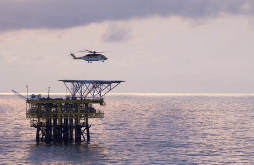Screenshot 2026-03-06 at 204224 - N9BO℠ | Global Underwater Services Ltd A helicopter approaches to land on the helipad of an offshore oil platform surrounded by calm ocean waters under a cloudy sky at dusk.