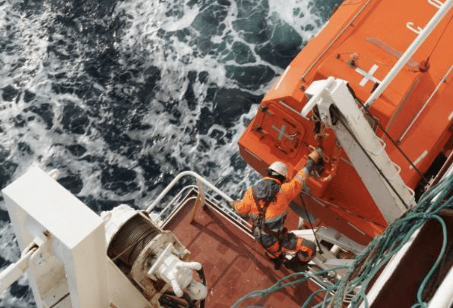 A worker in an orange safety suit and helmet stands on a ship’s deck next to an orange lifeboat, with ocean waves visible below. Ropes and safety equipment are nearby.