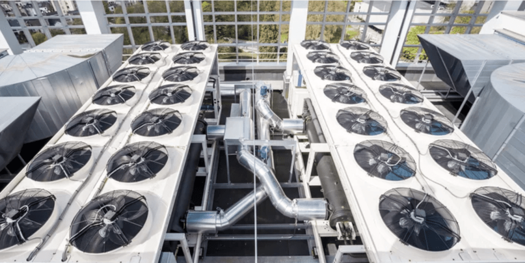Large industrial fans and ventilation ducts on the roof of a modern building, surrounded by glass windows and metal structures, with greenery visible outside.