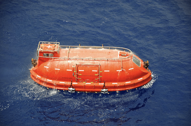 A bright orange enclosed lifeboat floats on calm, deep blue ocean water, viewed from above. The lifeboat’s hatch and safety rails are visible.
