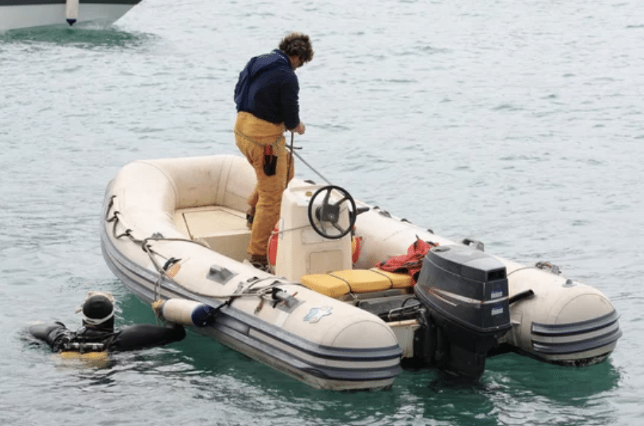 Screenshot 2026-03-06 at 225932 - N9BO℠ | Global Underwater Services Ltd A person in yellow trousers stands on an inflatable motorboat in the water, holding a rope. A scuba diver is partially submerged next to the boat’s side. The scene appears calm, with clear blue water.