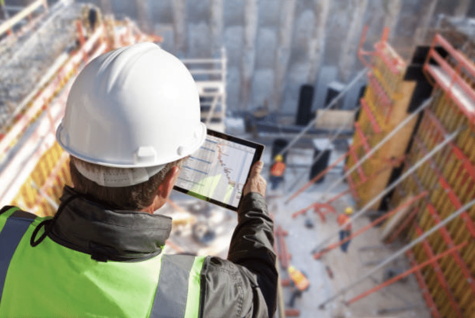 A construction worker wearing a white hard hat and yellow safety vest uses a tablet to review plans at a building site with scaffolding and other workers in the background.
