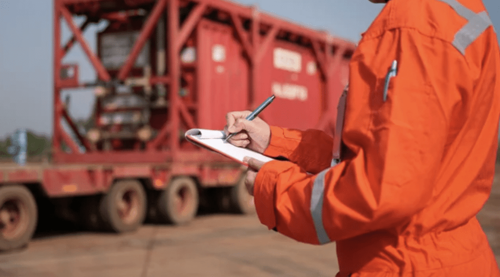 A person in an orange safety uniform writes on a clipboard whilst standing outdoors near large red industrial equipment on a flatbed trailer.