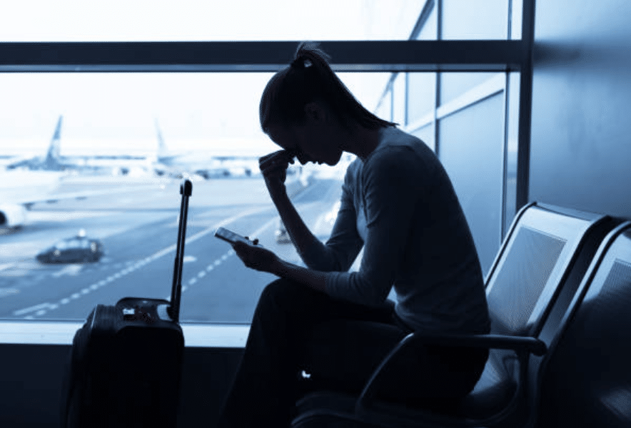 A woman sits in an airport terminal, holding her mobile and resting her head on her hand, appearing stressed or upset. A suitcase is beside her, and aeroplanes are visible through the large window in the background.