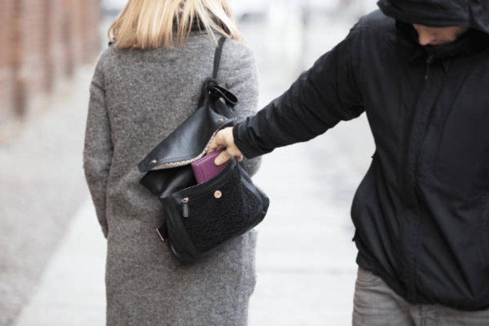 A person in a black hoodie is stealing a purple purse from the rucksack of a woman walking ahead, who is wearing a grey coat.