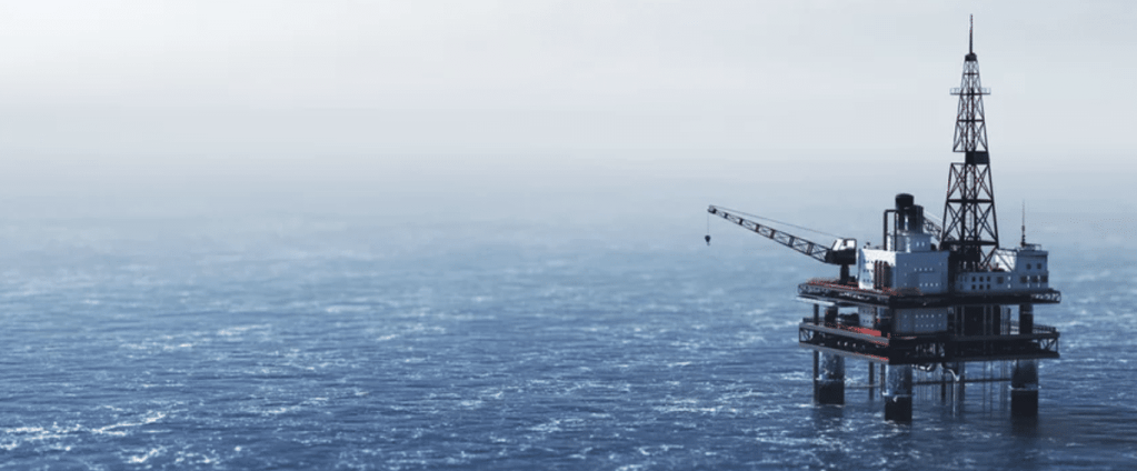 An offshore oil rig stands on pillars above the sea, surrounded by calm blue water and a hazy sky, with drilling and crane equipment visible on the platform.
