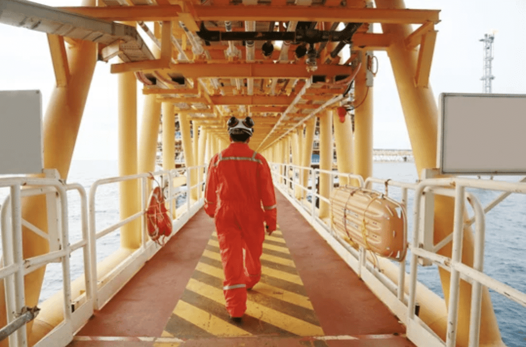 A worker in an orange safety suit and helmet walks along a yellow and orange metal platform on an offshore oil rig above the sea. Safety equipment and the sea are visible.