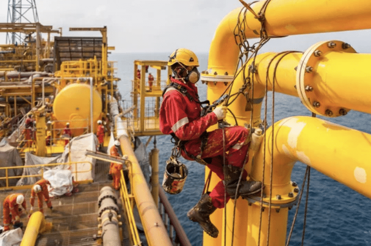 A worker in protective gear and harness performs maintenance on large yellow pipes at an offshore oil rig, with the sea and other workers visible in the background.