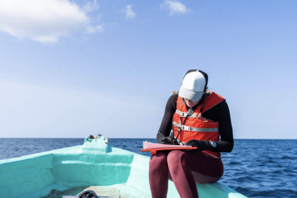 A person wearing a life jacket, cap, and gloves sits on a turquoise boat at sea, writing on a clipboard under a clear blue sky.