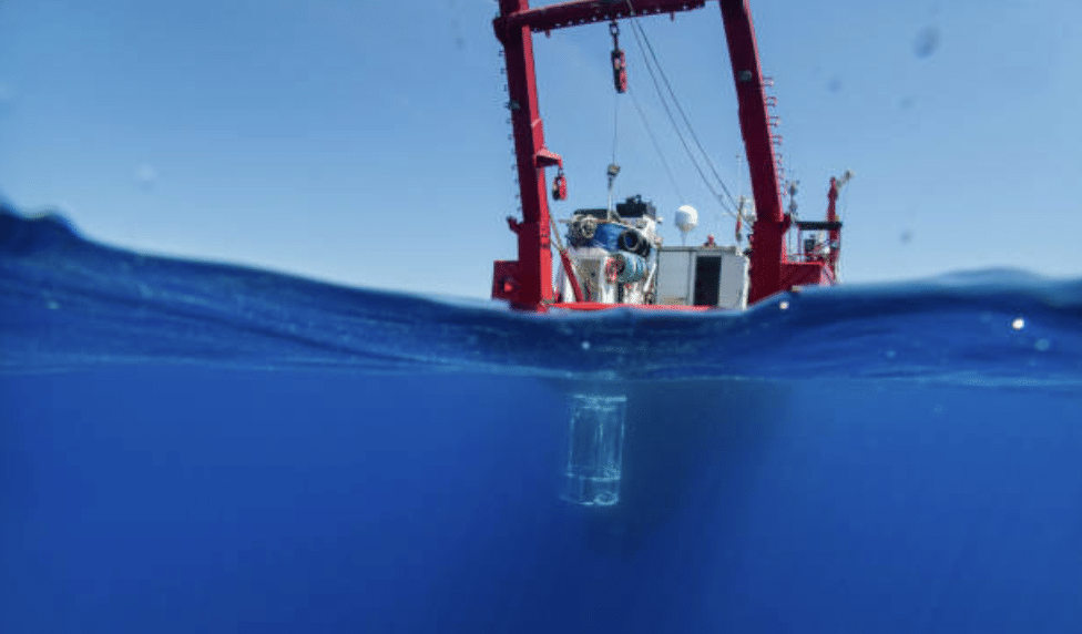 A red research vessel floats on the ocean, with scientific equipment partially submerged in clear blue water, viewed from a half-above, half-below perspective.