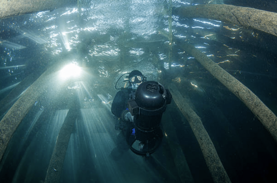Screenshot 2026-03-07 at 013827 - N9BO℠ | Global Underwater Services Ltd A scuba diver uses an underwater scooter to explore a submerged structure with wooden beams as sunlight filters through the water above.