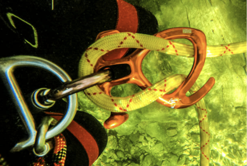 Screenshot 2026-03-07 at 014914 - N9BO℠ | Global Underwater Services Ltd Close-up of climbing equipment underwater, showing a yellow rope threaded through an orange belay device, attached to a silver karabiner. Submerged rocks and green-tinted water are visible in the background.