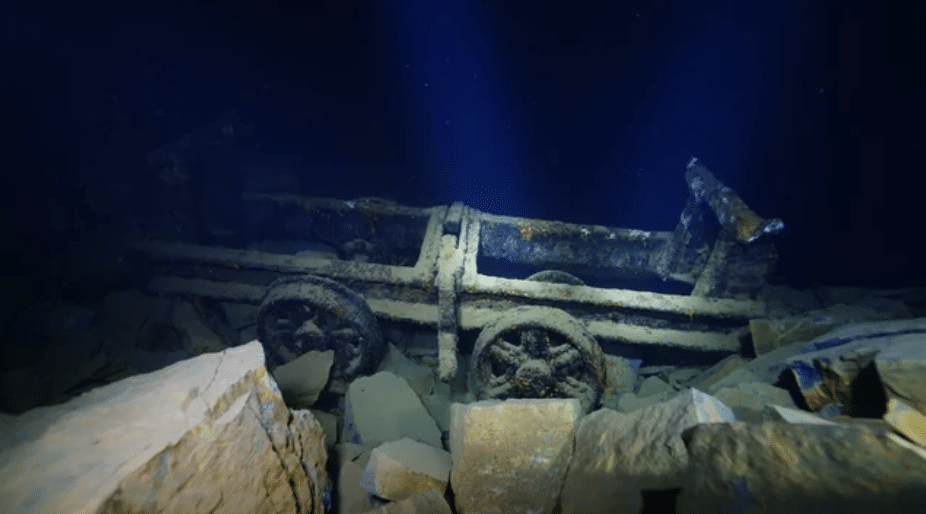 Screenshot 2026-03-07 at 020414 - N9BO℠ | Global Underwater Services Ltd An old, rusted mining trolley rests underwater on rocky debris, illuminated by beams of light filtering through the dark blue water.