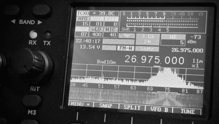 Black and white close-up of a radio transceiver display showing various frequency, signal, and tuning information, with dials and buttons visible on the left side of the device.