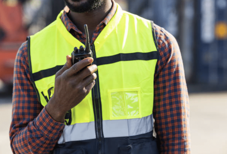 Screenshot 2026-03-14 at 115404 - N9BO℠ | Global Underwater Services Ltd A person wearing a yellow safety vest and checked shirt holds a walkie-talkie near their mouth, appearing to communicate. The background is blurred, suggesting an outdoor or industrial setting.