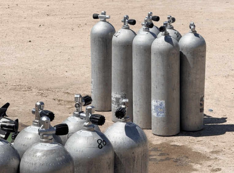 Screenshot 2026-03-14 at 120930 - N9BO℠ | Global Underwater Services Ltd A group of metal oxygen or scuba cylinders is arranged on a sandy, sunlit ground. Some cylinders are upright while others lie horizontally. The cylinders have visible valves and show signs of use and wear.
