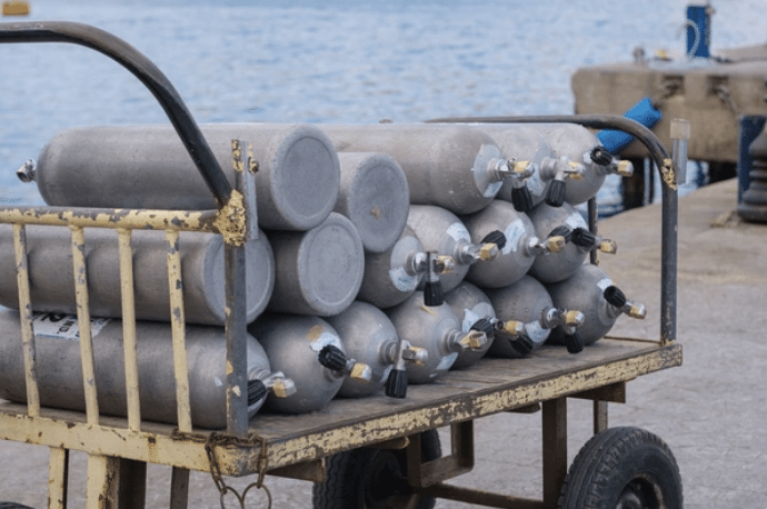 Screenshot 2026-03-14 at 121219 - N9BO℠ | Global Underwater Services Ltd A yellow metal trolley holds several stacked scuba diving air cylinders near a waterfront, with the sea and a concrete quay visible in the background.