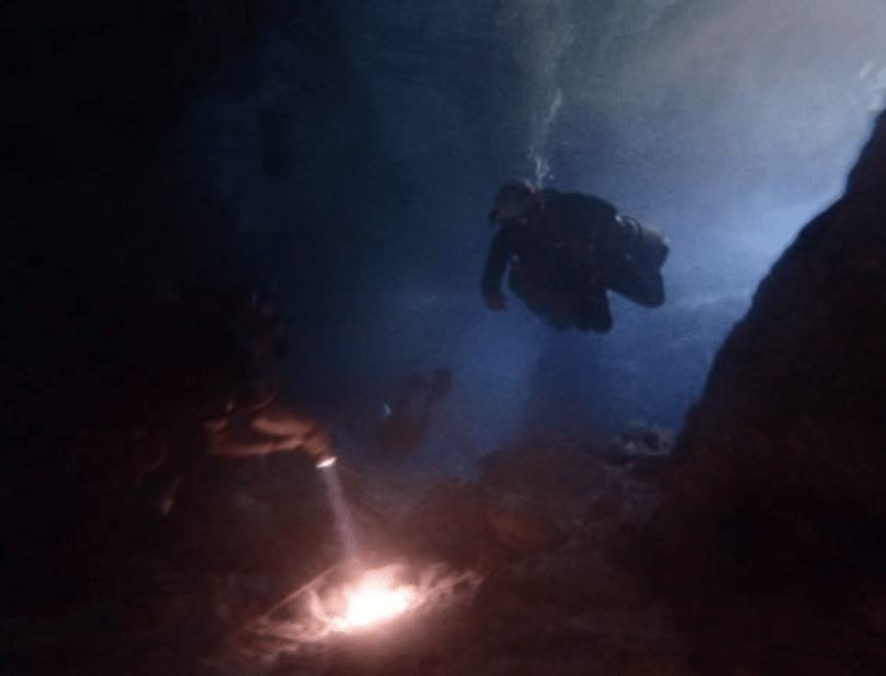 Screenshot 2026-03-14 at 132320 - N9BO℠ | Global Underwater Services Ltd Two scuba divers explore an underwater cave; one diver points a torch at the cave floor, illuminating the rocky terrain in the dim, blue water.