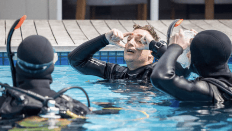 Three people in wetsuits and scuba gear are in a swimming pool. One person is demonstrating how to clear a scuba mask, whilst the other two watch attentively. The poolside and chairs are visible in the background.