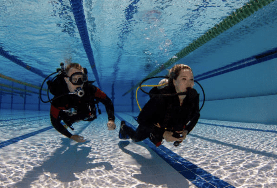 Screenshot 2026-03-14 at 133141 - N9BO℠ | Global Underwater Services Ltd Two people wearing scuba diving gear swim underwater in a clear, indoor swimming pool, following the lane lines along the bottom.