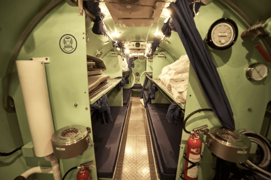 Screenshot 2026-03-14 at 140751 - N9BO℠ | Global Underwater Services Ltd The interior of a narrow, green-walled submarine or underwater chamber with bunk beds on each side, fire extinguishers, dials, and safety equipment, all under bright overhead lighting.