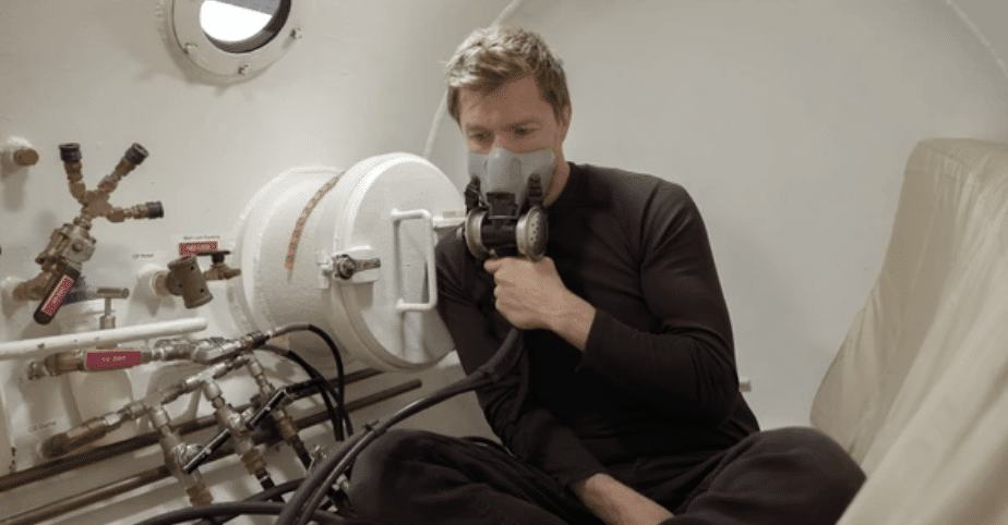 Screenshot 2026-03-14 at 140806 - N9BO℠ | Global Underwater Services Ltd A person sits inside a white chamber wearing a black outfit and a breathing mask connected to tubes and valves, suggesting they are undergoing a medical or scientific procedure.