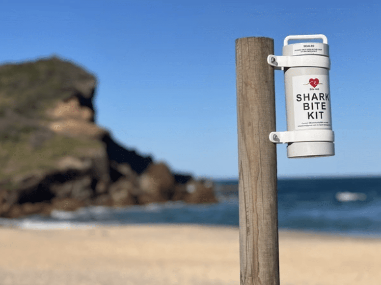 A white Shark Bite Kit is mounted on a wooden post at a beach, with sand, ocean waves, and a rocky outcrop visible in the background under a clear blue sky.