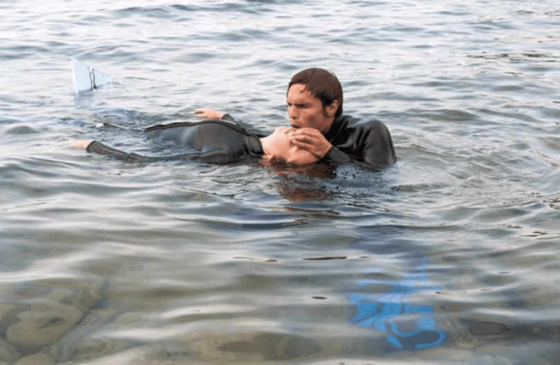 A person in a wetsuit gives mouth-to-mouth resuscitation to another person floating on their back in the water, both partially submerged, with a snorkel and flippers visible.