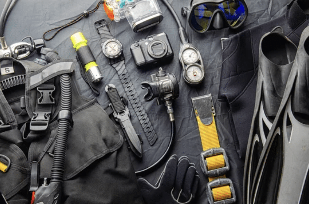 Screenshot 2026-03-14 at 152514 - N9BO℠ | Global Underwater Services Ltd A flat lay of scuba diving gear, including a diving mask, snorkel, regulator, gauges, underwater camera, torch, diving watch, gloves, fins, and a wetsuit, all arranged on a dark surface.