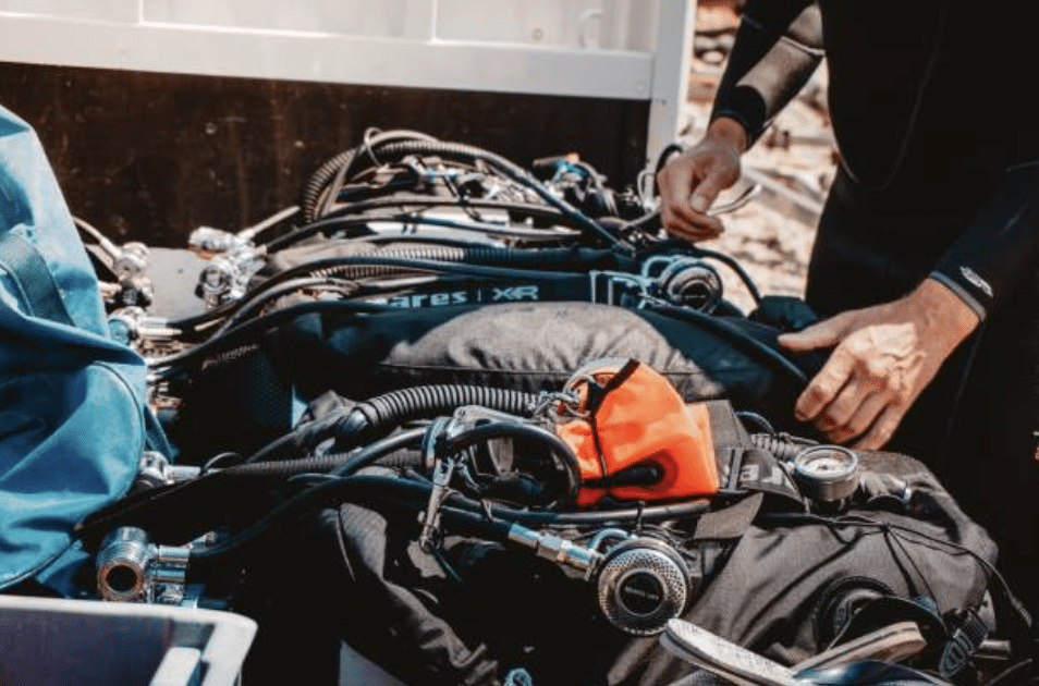 Screenshot 2026-03-14 at 152715 - N9BO℠ | Global Underwater Services Ltd A person wearing a wetsuit organises scuba diving equipment, including oxygen cylinders, gauges, and a bright orange safety marker, laid out on a surface outdoors.