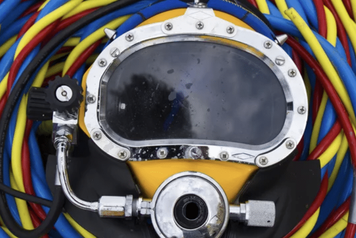 A close-up of a yellow and chrome deep-sea diving helmet, surrounded by coiled blue, red, and yellow hoses. The helmet's glass visor and metal fittings are visible.
