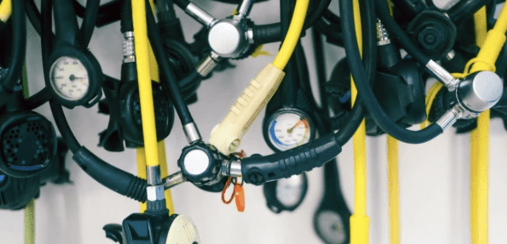 Screenshot 2026-03-14 at 152916 - N9BO℠ | Global Underwater Services Ltd A close-up of several scuba diving regulators and hoses with attached pressure gauges, mostly black and yellow, hanging together in a cluster.