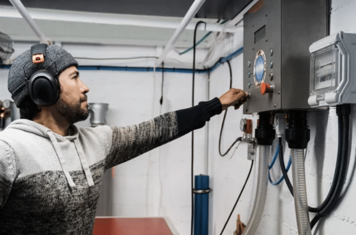 Screenshot 2026-03-14 at 153659 - N9BO℠ | Global Underwater Services Ltd A man wearing a knitted jumper, beanie, and ear defenders is operating a control panel with various buttons and a circular display in an industrial or mechanical room.