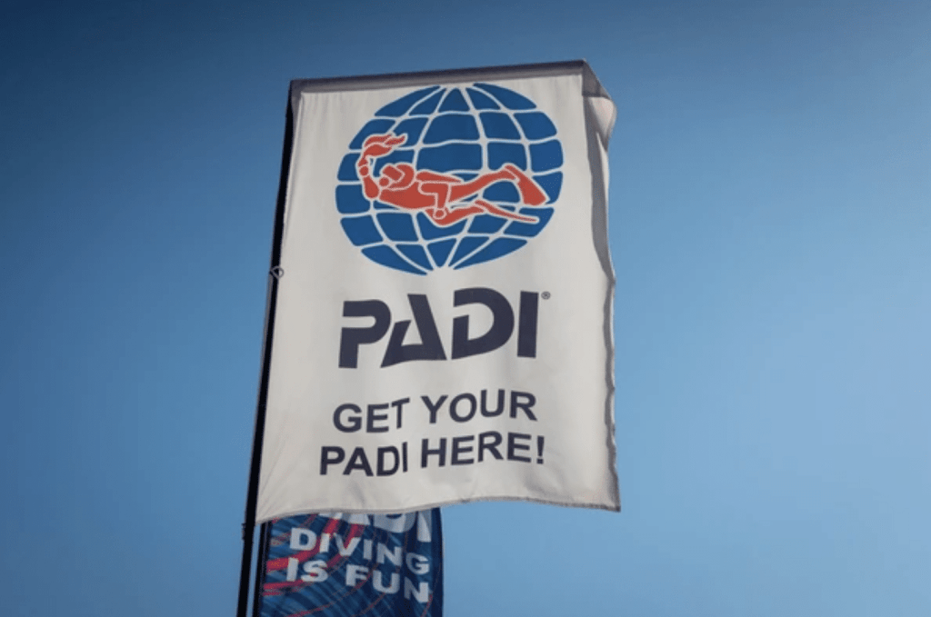 A white flag with the PADI logo, showing a red scuba diver and a globe, and the text PADI GET YOUR PADI HERE! waving against a clear blue sky.
