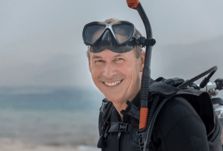 Screenshot 2026-03-14 at 160513 - N9BO℠ | Global Underwater Services Ltd A smiling man in a wetsuit and scuba gear stands outdoors near the water, wearing a snorkel mask on his forehead.