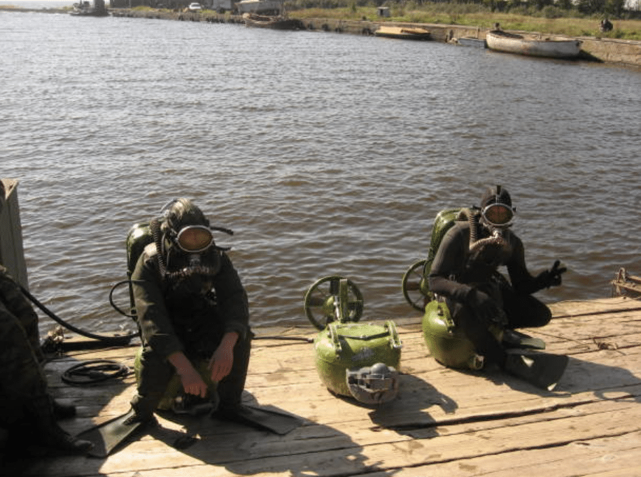 Screenshot 2026-03-14 at 161426 - N9BO℠ | Global Underwater Services Ltd Two divers in vintage diving suits with helmets and oxygen tanks sit on a wooden jetty by a river. Diving equipment and flippers are lying nearby. The water and some boats are visible in the background.