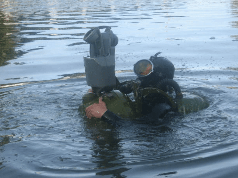Screenshot 2026-03-14 at 161436 - N9BO℠ | Global Underwater Services Ltd A diver in full kit, including breathing apparatus and mask, is partially submerged in water, holding a large piece of equipment above the surface. The surroundings are calm with gentle ripples on the water.