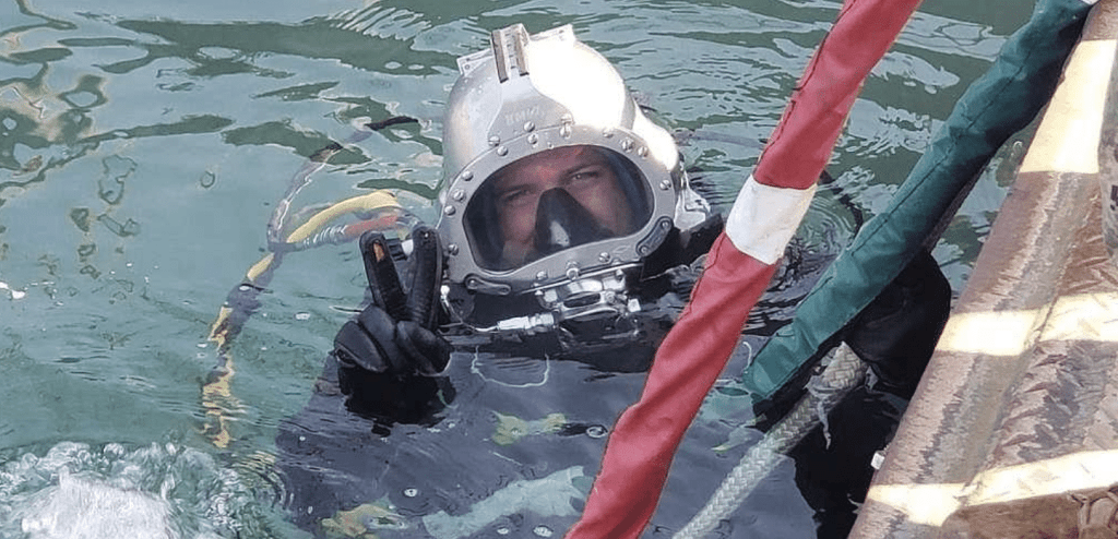 A diver in a heavy-duty helmet and suit floats in greenish water, holding onto a rope with one hand and making a peace sign with the other. Cables and a striped pole are visible beside the diver.