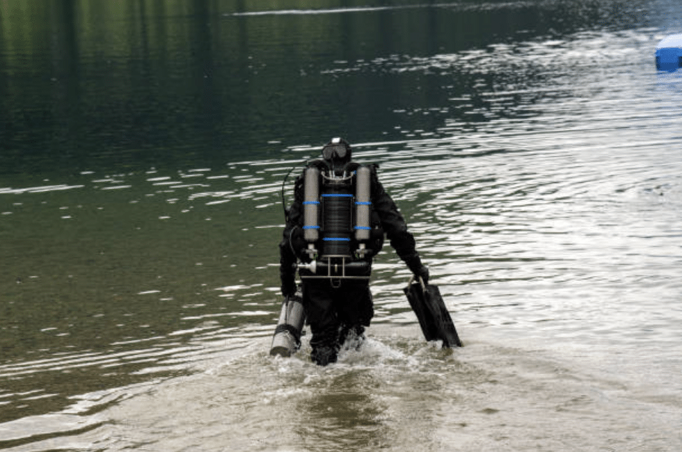 Screenshot 2026-03-14 at 181953 - N9BO℠ | Global Underwater Services Ltd A scuba diver wearing a black wetsuit and carrying diving equipment walks into calm, shallow water from the shore, with ripples forming around their legs.
