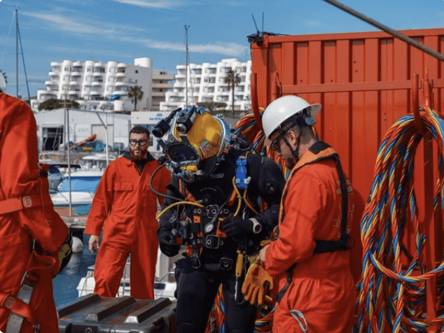 Four workers in orange overalls, two with white helmets, assist a person in a black diving suit and helmet near bright coiled cables at a quay with white buildings and boats in the background.