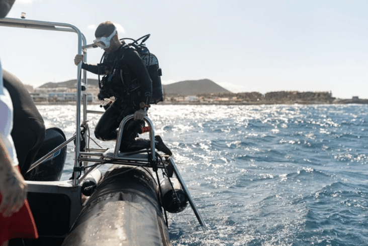 Screenshot 2026-03-14 at 185345 - N9BO℠ | Global Underwater Services Ltd A scuba diver wearing full kit prepares to enter the water from the side of a boat, with a coastal landscape and blue, choppy sea in the background.