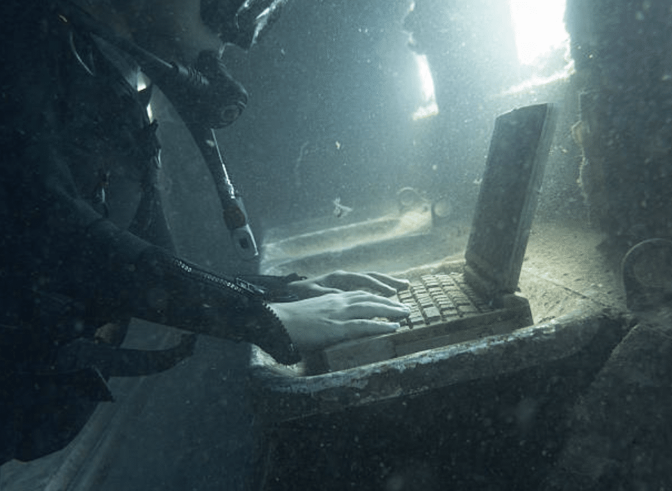 A scuba diver in full kit types on an old, dust-covered laptop inside a submerged, sunken structure with light shining through a nearby window.