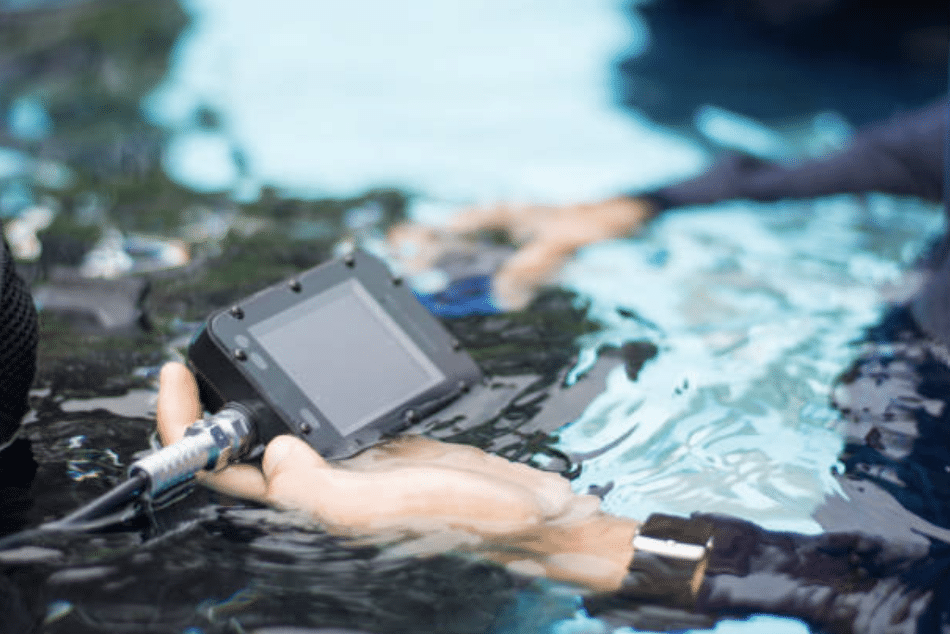 A person holds an electronic device partially submerged in water, with the screen and cables visible. Another hand and blurred background suggest a pool or aquatic setting.