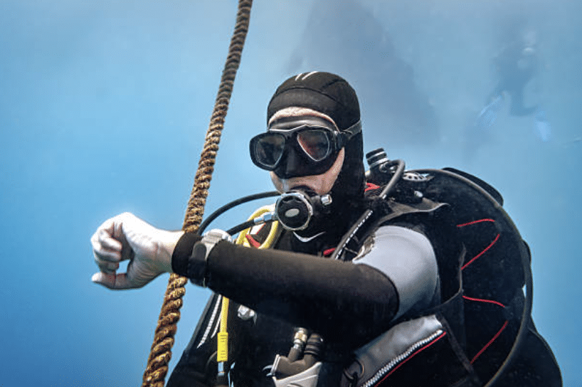 A scuba diver in full kit holds onto a rope underwater and checks their wristwatch or dive computer, with another diver visible in the background against a blue aquatic environment.