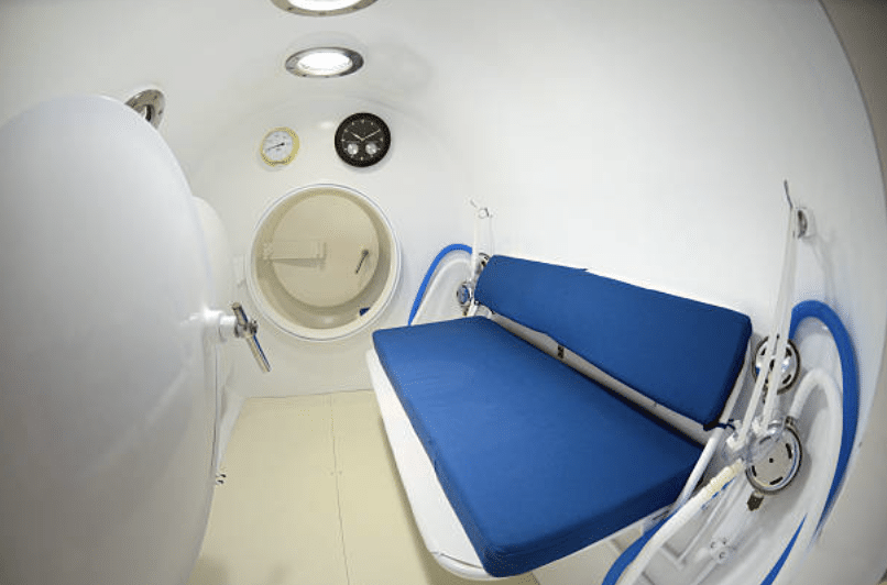 Screenshot 2026-03-14 at 230654 - N9BO℠ | Global Underwater Services Ltd Interior of a hyperbaric oxygen chamber with a blue padded bench, white walls, two round dials, and a black clock above a sealed round door, all under bright ceiling lights.