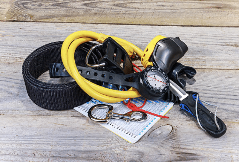 Screenshot 2026-03-14 at 230739 - N9BO℠ | Global Underwater Services Ltd A pile of scuba diving equipment, including a yellow regulator hose, a diving computer with compass, a black nylon strap, a logbook, a whistle, a dive slate, and a metal clip, all on a wooden surface.
