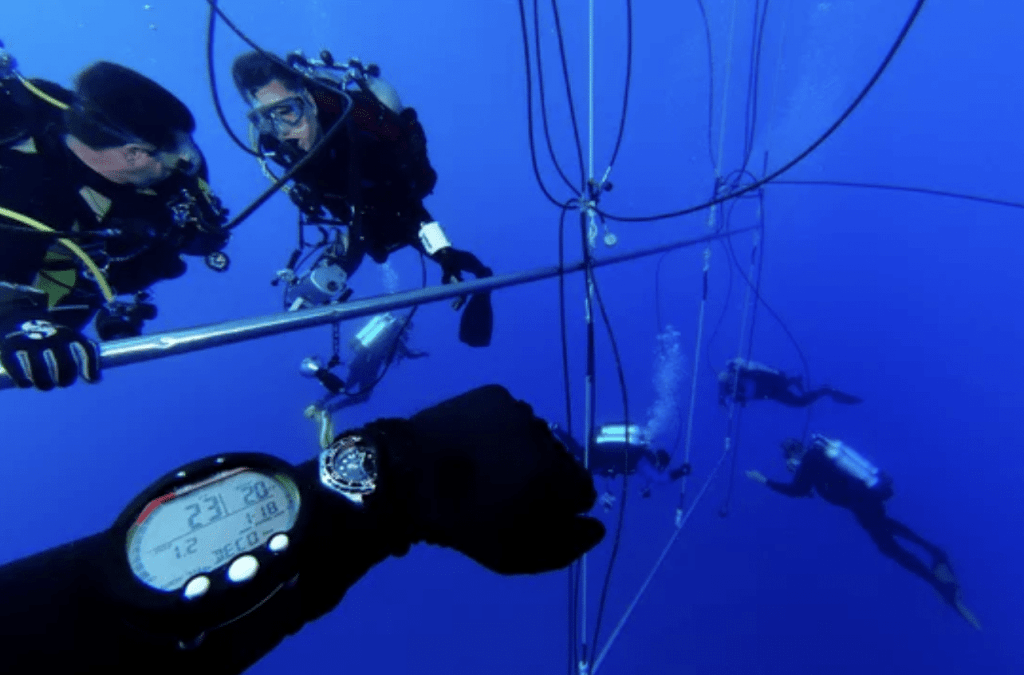 Three scuba divers are underwater near a metal rod and wires, with a diver’s wrist showing a digital dive computer in the foreground. Additional divers are swimming in the blue water in the background.