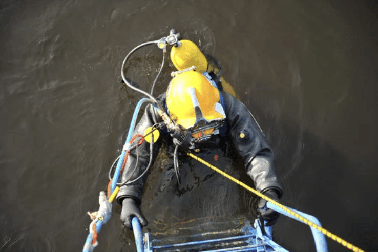 A diver in a yellow helmet and black wetsuit climbs a ladder out of dark water whilst holding a rope, with various hoses and cables attached to their suit.
