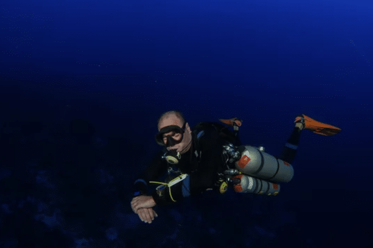 A scuba diver wearing a black wetsuit, orange fins, and multiple oxygen cylinders swims underwater in deep blue ocean, surrounded by dark water with minimal visible marine life.
