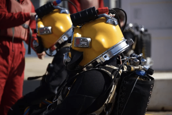 Screenshot 2026-03-14 at 233548 - N9BO℠ | Global Underwater Services Ltd Two people wearing yellow diving helmets and black wetsuits sit side by side, equipped with breathing apparatus. Another person in a red suit stands nearby, partially visible. The setting appears to be outdoors.
